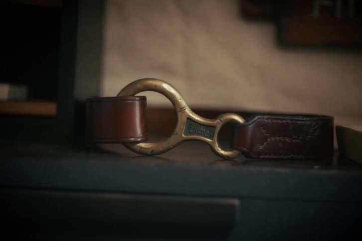 A close-up still life of a classic brown leather belt with a brass buckle resting on a dark surface