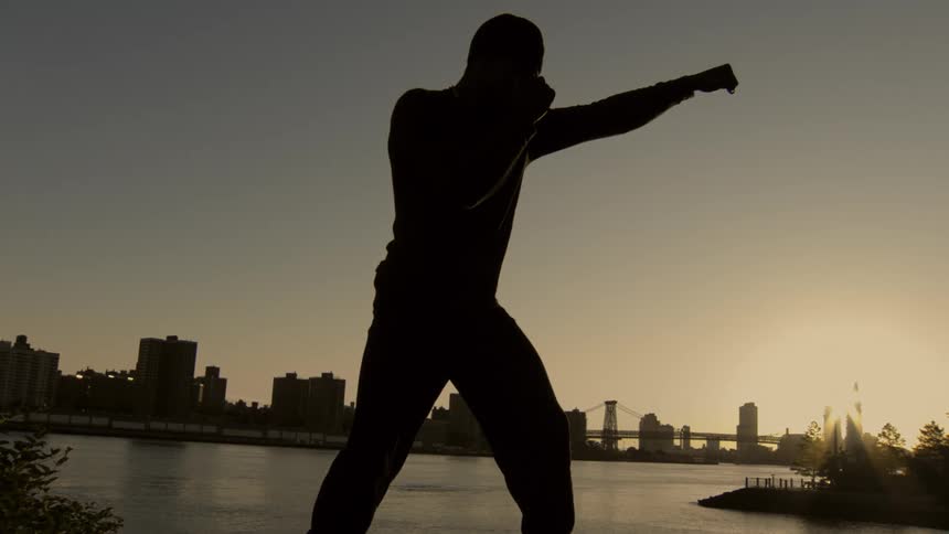 A silhouette of a man practices boxing in slow motion against a city skyline at sunset