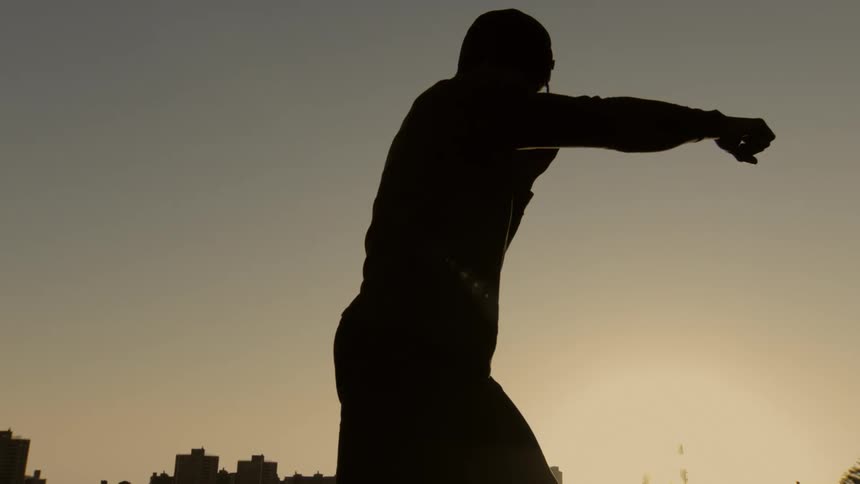 A slow-motion cinematic silhouette shows a determined boxer practicing his punch against a city skyline at sunset
