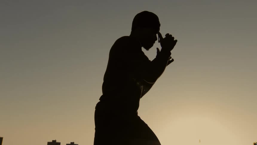 A silhouette of a man holding a defensive boxing pose in slow motion against the evening sky