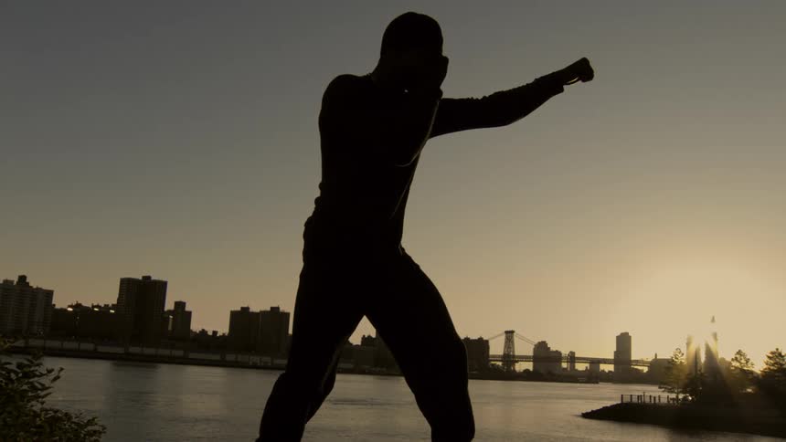 A silhouetted man shadow boxes in slow motion, throwing a punch against the backdrop of a city skyline at sunset