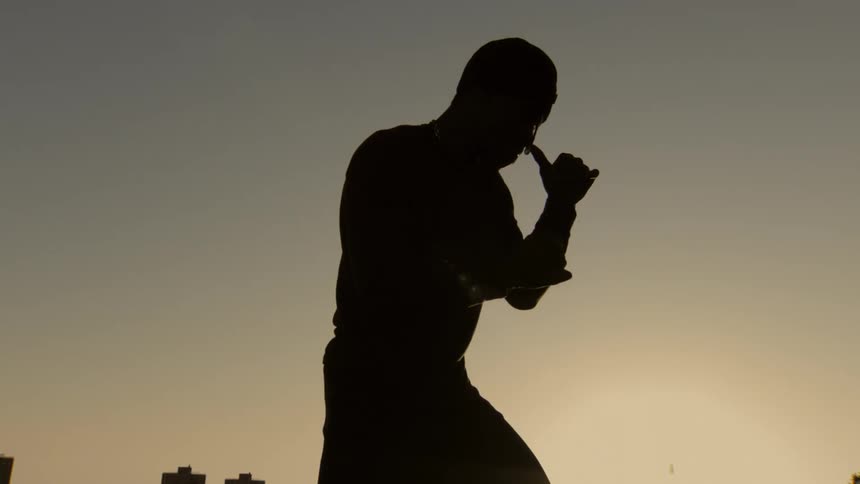 A silhouetted boxer holds a fighting stance in slow motion against the sunset sky above a city