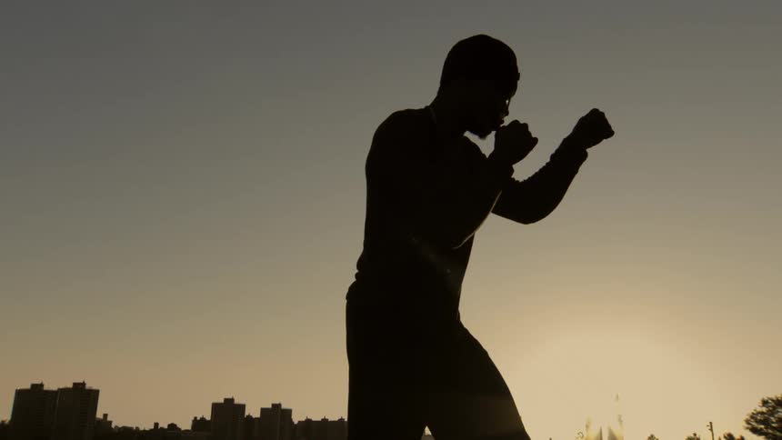A silhouetted man shadowboxes in slow motion against a golden city skyline at sunset