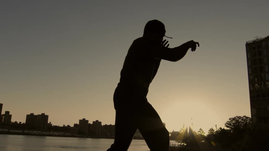 A silhouetted man practices martial arts in slow motion by an urban waterfront at sunset