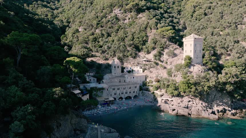 A beautiful aerial view reveals the historic San Fruttuoso Abbey and its secluded beach nestled in a cove on the Italian Riviera, with people swimming in the turquoise water