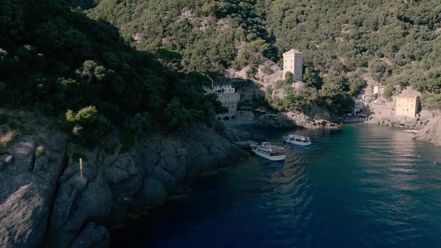 Aerial view flying over sailboats in a pristine blue bay towards the historic San Fruttuoso Abbey on the Italian Riviera