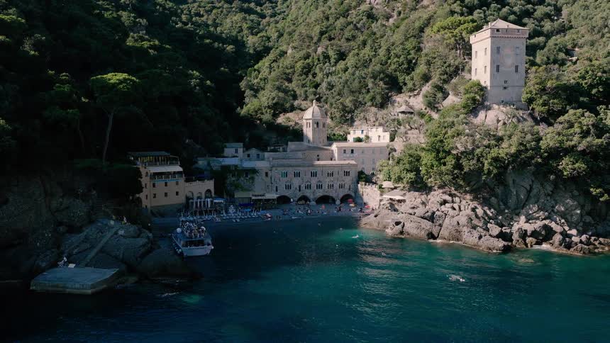 An aerial view pulls back from the historic San Fruttuoso Abbey to reveal a stunning secluded beach cove on the Italian Riviera, bustling with sunbathers and swimmers