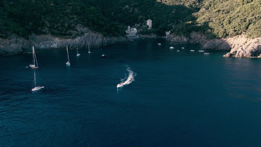 An aerial view of a motorboat cruising across a stunning blue bay with sailboats anchored near a secluded cove on a lush coastline