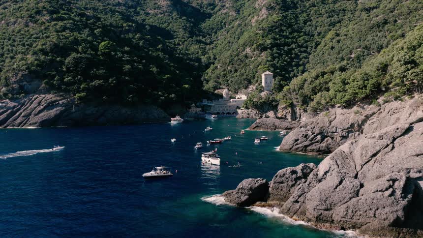 An aerial pullback shot reveals the stunning San Fruttuoso bay, where boats are anchored and people swim in the beautiful blue waters