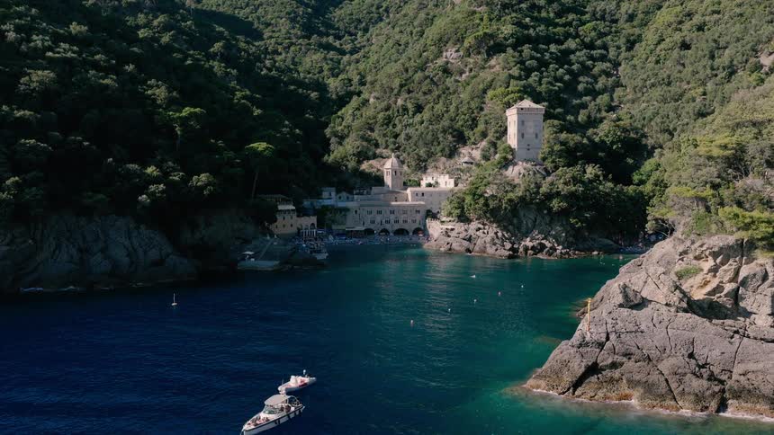 An aerial view of the historic San Fruttuoso Abbey with its iconic watchtower, nestled in a secluded cove with boats floating on the tranquil blue sea of the Italian Riviera