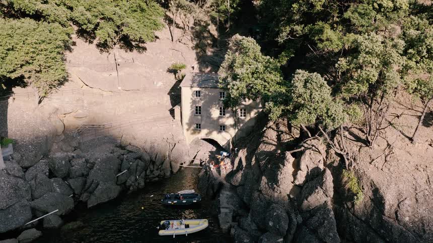 Aerial shot pulling up to reveal a unique historic building nestled into a rocky cove with small boats moored in the calm water below
