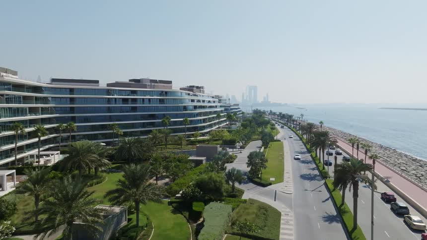 A scenic aerial shot flies over a luxurious oceanfront resort in Dubai, showing the coastal road and distant city skyline on a sunny day