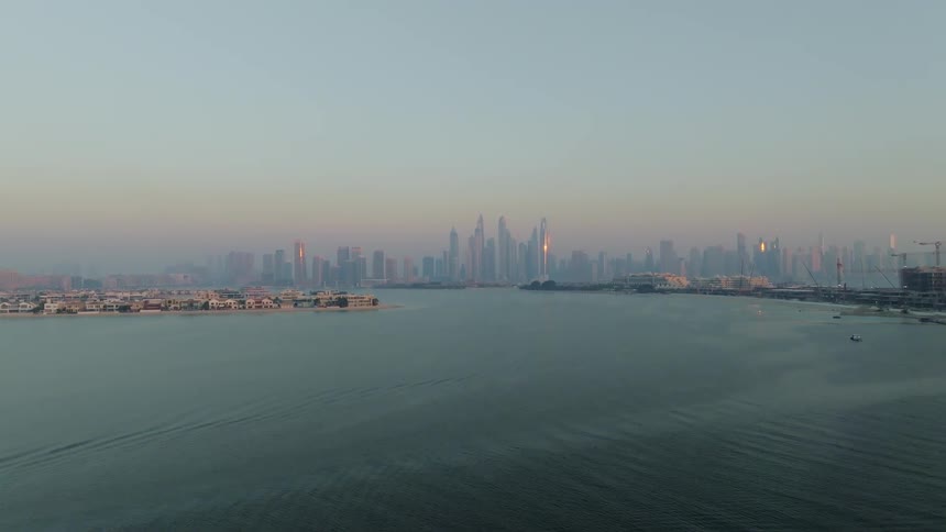 A static aerial view of the modern Dubai cityscape across the ocean during a hazy sunrise