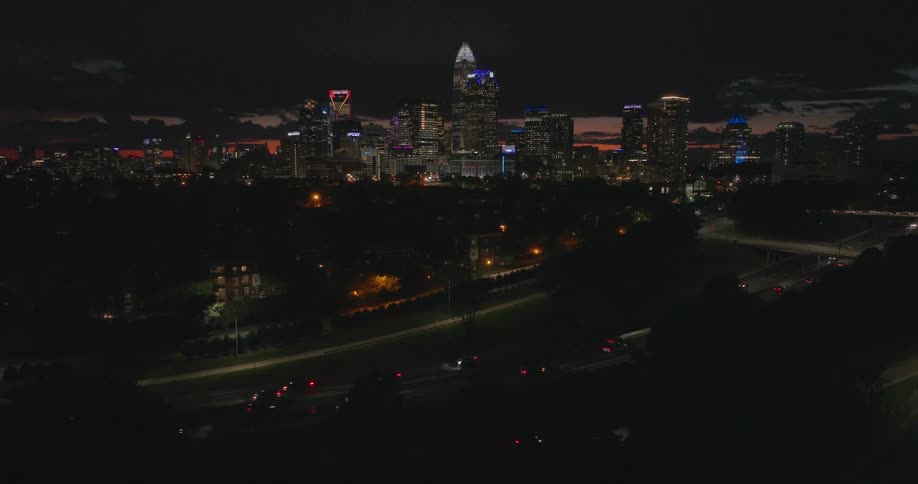 An aerial view pulls back from a busy highway at night to reveal the glowing Charlotte, North Carolina city skyline at dusk
