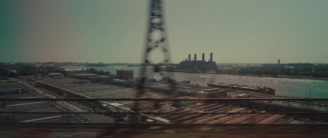 A cinematic tracking shot from a moving train or vehicle shows an expansive industrial landscape with a power plant and shipping port along a river