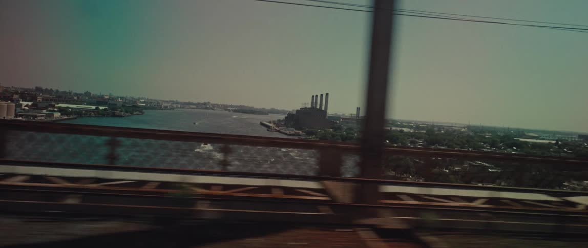A side-view shot from a moving train crossing a large bridge, showing an industrial cityscape and river