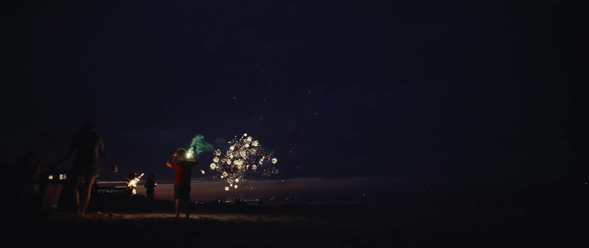 A family on the beach at night enjoys a spectacular fireworks show while playing with sparklers in slow motion