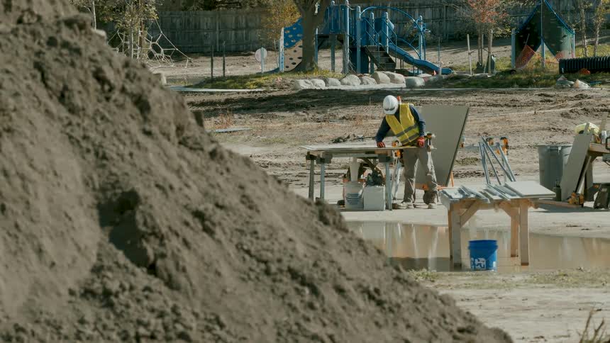 A construction worker saws material at a muddy work site with a large dirt pile in the foreground and a playground behind him