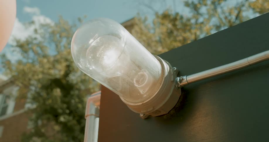 A close-up of an industrial outdoor light fixture with a glass cover, glowing during the daytime with trees and a building in the background