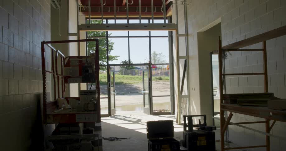 A slow zoom out reveals the interior of an unfinished building hallway under construction, looking out a large glass doorway to a sunny playground