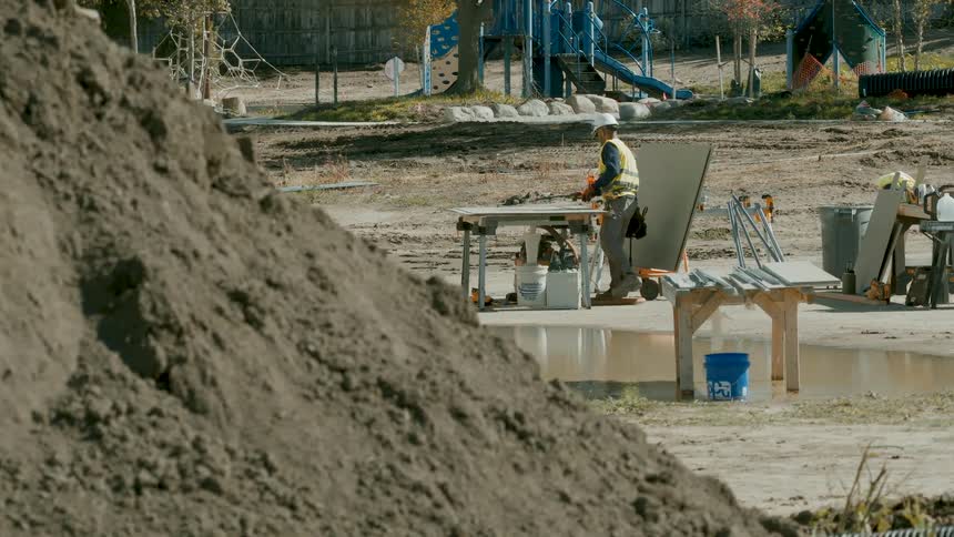 A slow pan reveals a construction worker in a hard hat working at a table behind a large dirt pile on a job site
