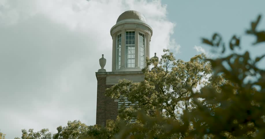 A stately brick building's cupola stands tall against a partly cloudy sky, framed by lush green trees