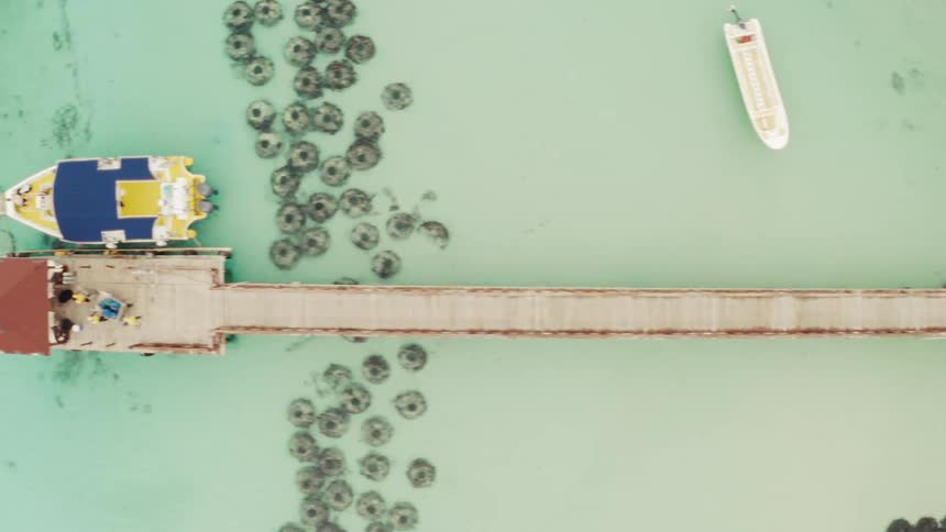 Aerial top-down view follows a long wooden pier from a sandy tropical beach with kayaks out into the clear turquoise ocean