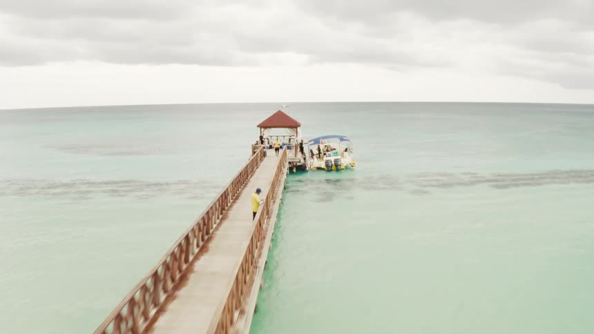 Aerial drone flies over a beautiful beach and a long wooden pier where people are getting on tour boats in the turquoise Caribbean sea