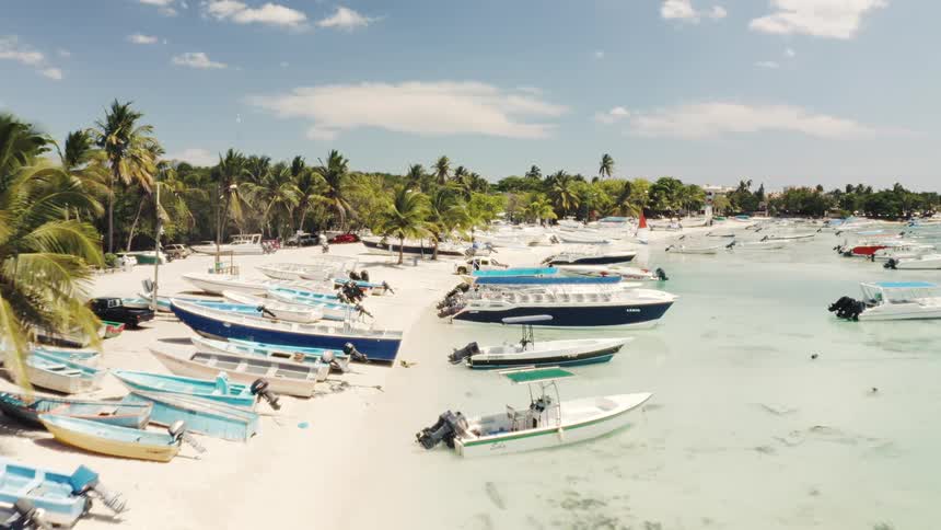 Slow-moving aerial shot of a sunny tropical beach, showcasing numerous tour boats and fishing boats parked on the white sand and floating in the shallow turquoise sea