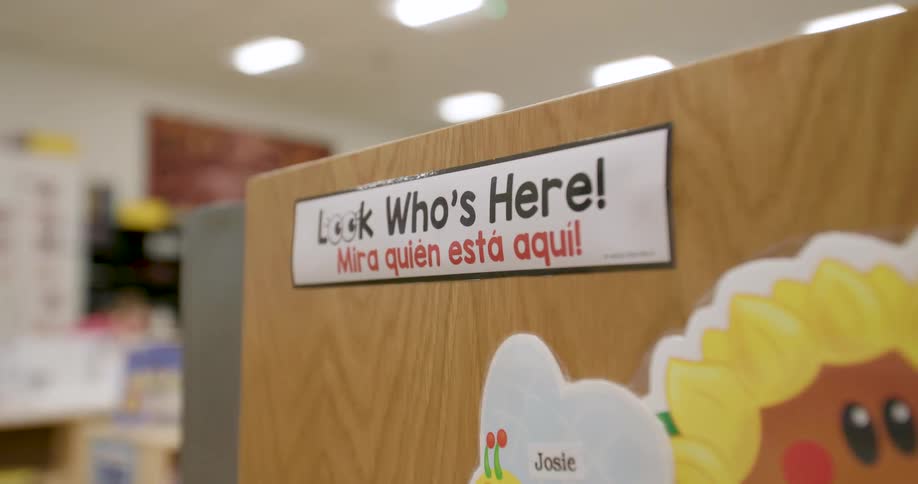 A close-up shot of a bilingual English and Spanish 'Look Who's Here!' sign in a brightly lit elementary school classroom