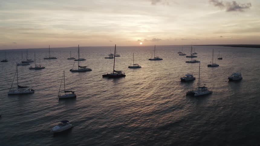 A stunning aerial view captures dozens of sailboats and catamarans floating peacefully in a tropical bay during a golden sunset