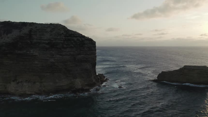 A dramatic aerial view pans across a large rock formation in the vast ocean as the sun sets on the horizon