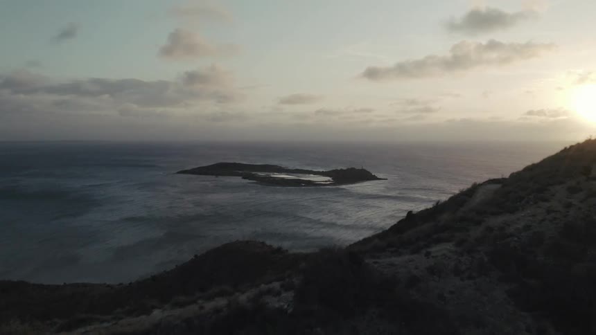 An aerial shot flies over a silhouetted mountain ridge revealing a remote island in the ocean at sunset