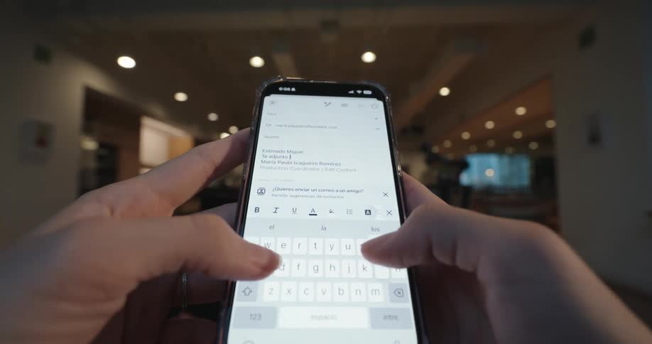 A close-up, first-person view of a person typing an email in Spanish on their smartphone in a warmly lit indoor setting