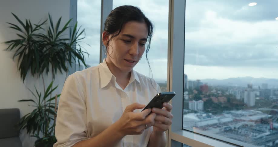 A young businesswoman smiles while texting on her smartphone in a modern high-rise office with a city view