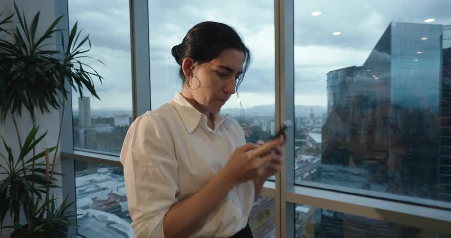 A businesswoman smiles while using her smartphone by the window of a modern high-rise office overlooking the city