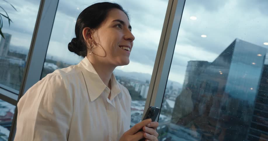 A happy businesswoman texts on her smartphone while standing by the window of her high-rise office overlooking the city