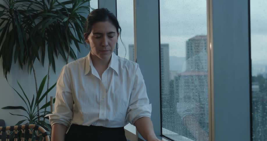 A stressed businesswoman pauses from her phone, taking a deep breath to find a moment of calm by a rainy window overlooking the city