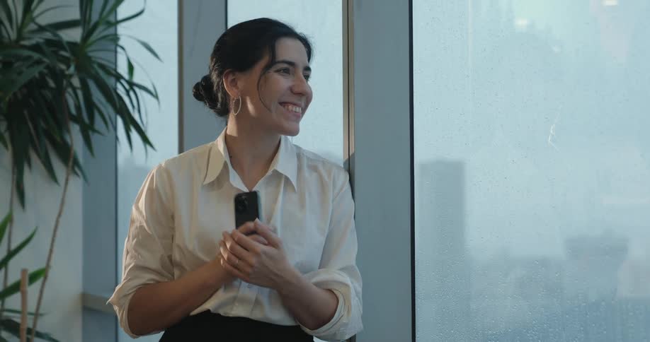 A professional woman in a white shirt smiles with delight after checking her phone while standing by a large, rainy window in her office
