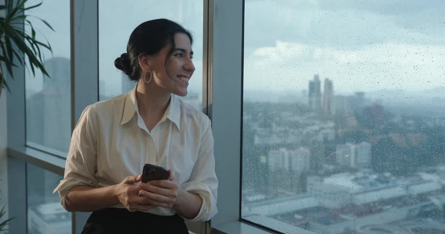A cheerful businesswoman gets a happy message on her phone and joyfully looks out the large office window at the rainy cityscape
