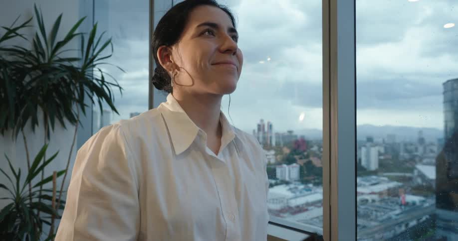A professional woman in a modern office smiles with satisfaction as she pauses from her phone to gaze at the sprawling city view