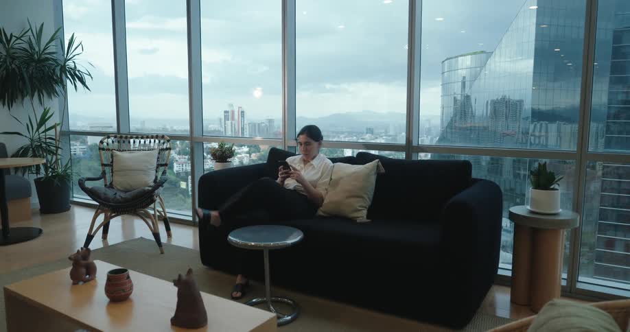 A woman in business attire walks over to a sofa and relaxes while using her smartphone in a contemporary office lounge with a panoramic city view