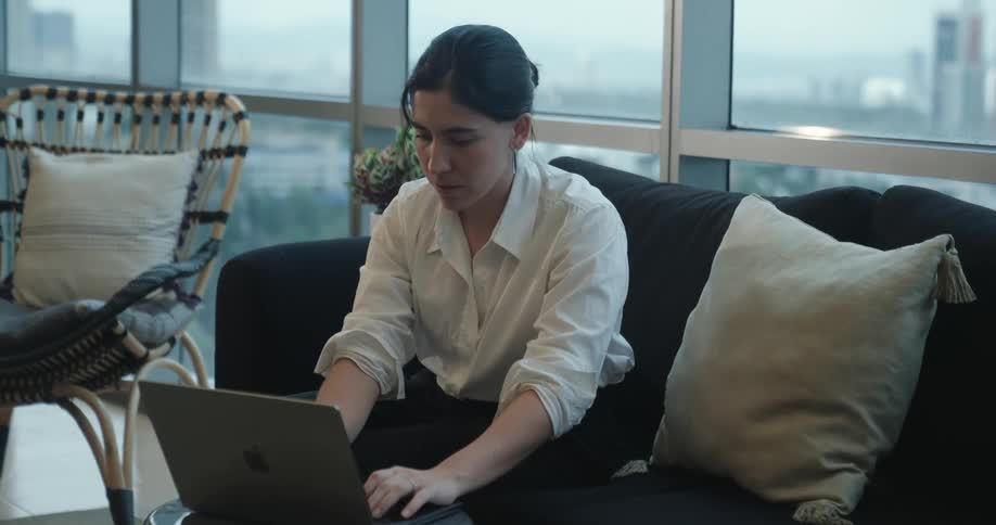 A young woman smiles while working on her laptop from the comfort of a sofa in her modern city apartment