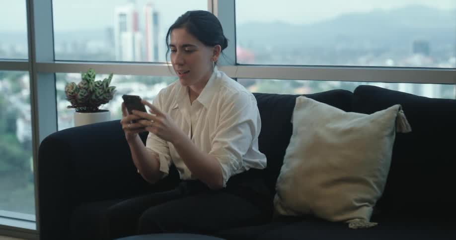 A professional woman sits down on a sofa in a modern high-rise office and smiles while using her smartphone