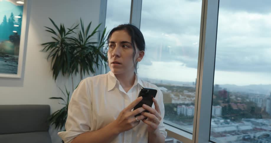 A businesswoman's expression shifts from concentration to joy as she uses her phone in a modern office overlooking the city at dusk
