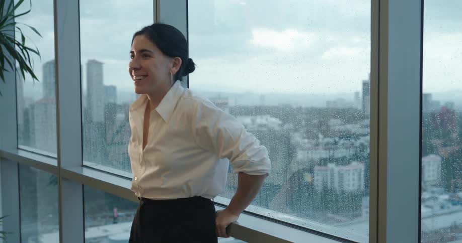 A happy young businesswoman smiles while looking out a high-rise office window at the rainy city below