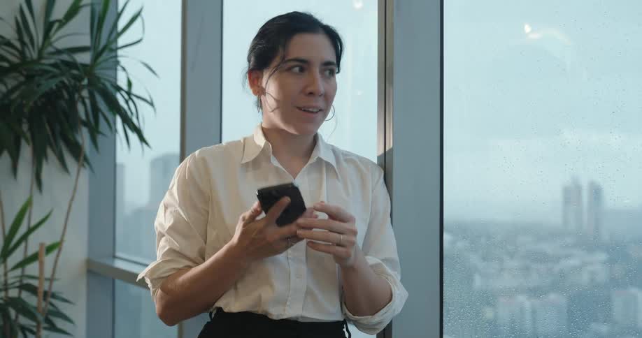 A happy businesswoman standing by the window of her high-rise office smiles after receiving good news on her phone