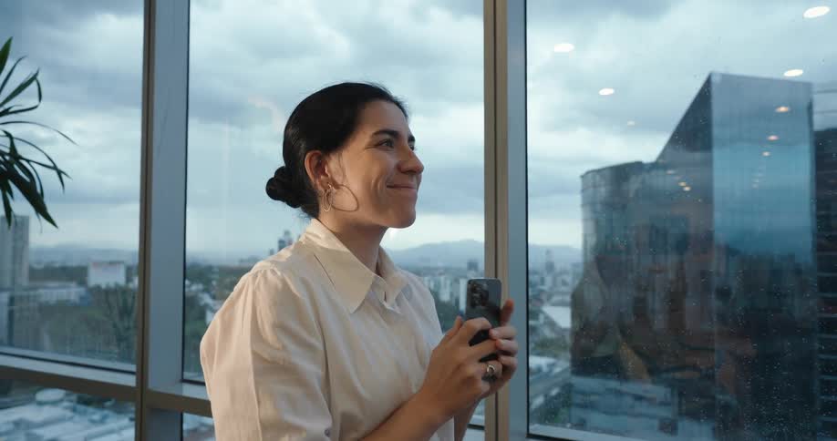 A happy businesswoman holds her phone while smiling and looking out a large office window at the city skyline on a cloudy day