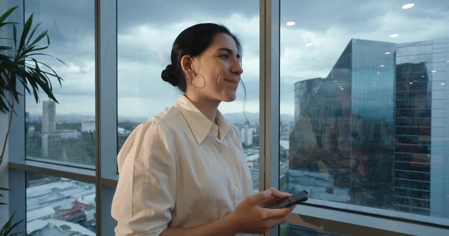 A businesswoman takes a break from her phone to smile and enjoy the expansive city view from her modern high-rise office