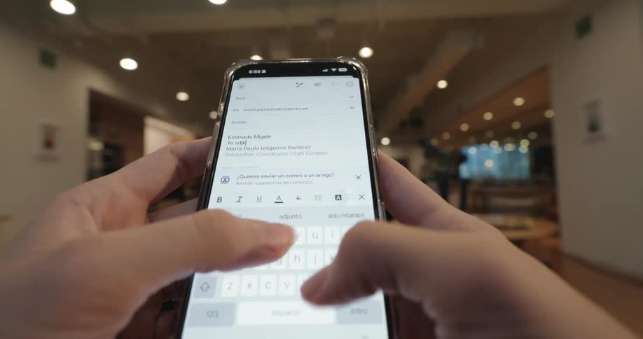 A first-person perspective of hands typing an email in Spanish on a smartphone in an office setting